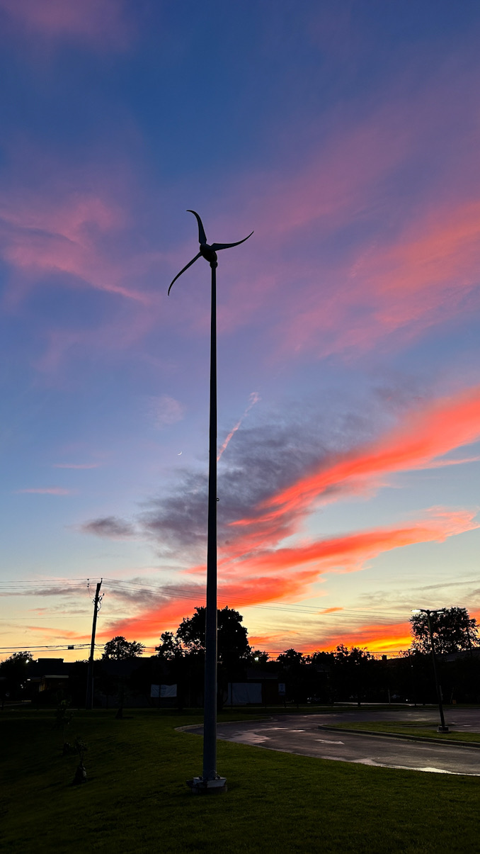 Wider angle photo captured at 8:42 p.m. Saturday, June 8, 2024, showing a Skystream 3.7 Wind Turbine at Thomas Middle School capable of generating power from wind, with a waxing crescent moon and sunset in the background (CARDINAL NEWS)
